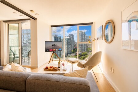 Bright living room with a city view, gray sofa, and a coffee table topped with snacks and wine; TV on a tripod stand faces the seating area.