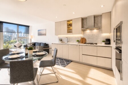 Open-plan kitchen and dining area in a bright apartment, glass dining table, grey chairs, marble backsplash, and city view.