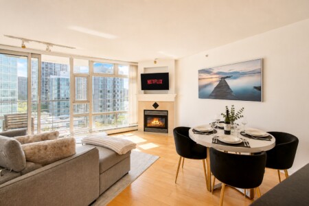 Bright urban living room with floor-to-ceiling windows, gray sofa, and a fireplace; Netflix on a wall-mounted TV above the mantel.