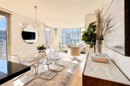 Sunlit open-plan dining area with a marble table, clear acrylic chairs, and a curved living room by large city-view windows.