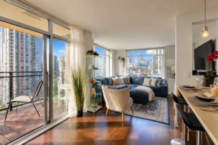 Bright living room with a blue sectional and beige chairs, facing large windows showing a city skyline; balcony on the left and decorative shelves nearby.