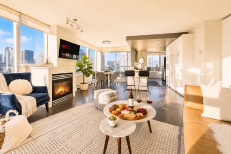 Bright open-concept living room with a fireplace, TV, and floor-to-ceiling windows overlooking a cityscape; blue sofa and beige seating with a cheese board on a marble coffee table.