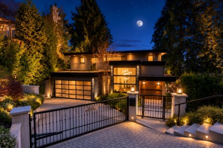 Night view of a modern two‑story villa with warm interior lights, gated driveway, and a full moon in a starry sky.