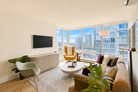 Sunlit modern living room with floor-to-ceiling windows offering a city skyline, beige sofa, orange swivel chair, and round white coffee table with plants nearby.