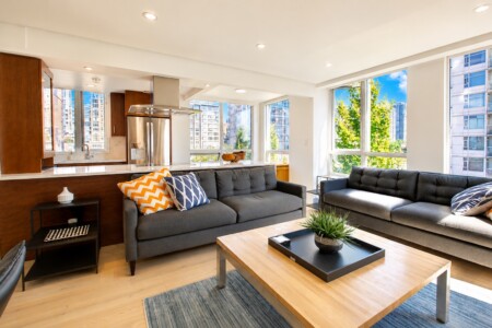 Bright open-concept living room with gray sofas, a wooden coffee table, and large windows showing city skyline.