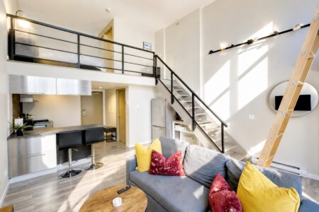 Open-plan loft with stainless steel kitchen, island seating for two, and a gray sofa with yellow and red cushions; staircase to a mezzanine.