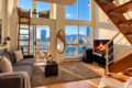 Modern open-plan living room with a gray sofa, rug, and a fireplace, overlooking a city skyline through large windows.