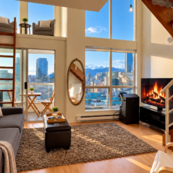 Modern open-plan living room with a gray sofa, rug, and a fireplace, overlooking a city skyline through large windows.