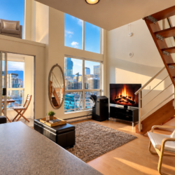 Modern loft living room with large city-view windows, a wooden staircase, and a fireplace beside a TV.