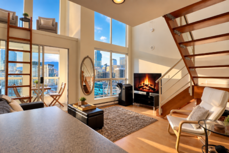 Modern loft living room with large city-view windows, a wooden staircase, and a fireplace beside a TV.