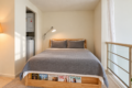 Wooden-framed bed with gray bedding and two matching pillows, bedside lamp overhead, magazines visible at the footboard. A laundry alcove is to the left; glass balcony railing and sheer curtain to the right.
