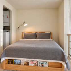 Wooden-framed bed with gray bedding and two matching pillows, bedside lamp overhead, magazines visible at the footboard. A laundry alcove is to the left; glass balcony railing and sheer curtain to the right.