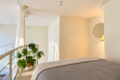 Bright, minimal-bedroom loft with a bed in the foreground, beige walls, and a round wall mirror with a warm lamp nearby.