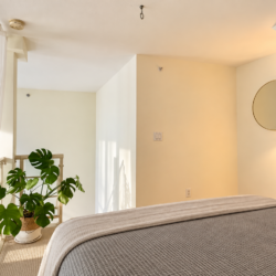 Bright, minimal-bedroom loft with a bed in the foreground, beige walls, and a round wall mirror with a warm lamp nearby.