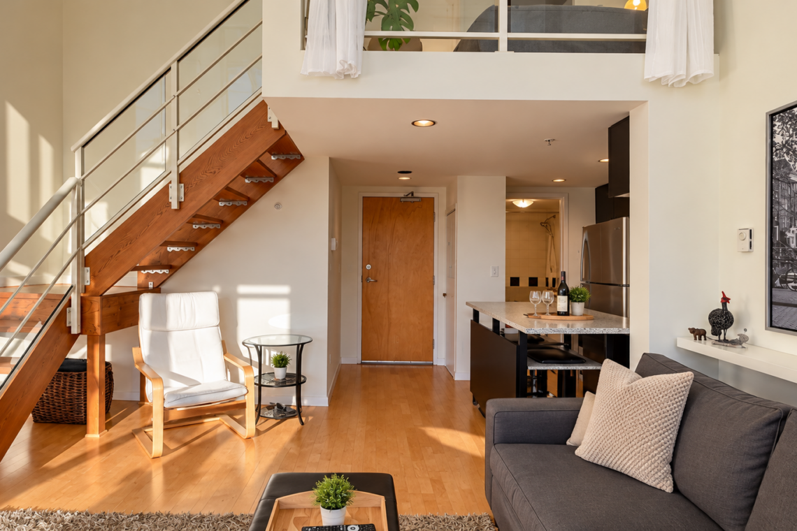 Open-plan living space with a wooden staircase, white lounge chair, glass side table, gray sofa, and a kitchen island.