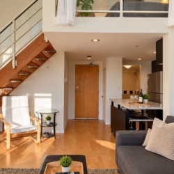 Open-plan living space with a wooden staircase, white lounge chair, glass side table, gray sofa, and a kitchen island.