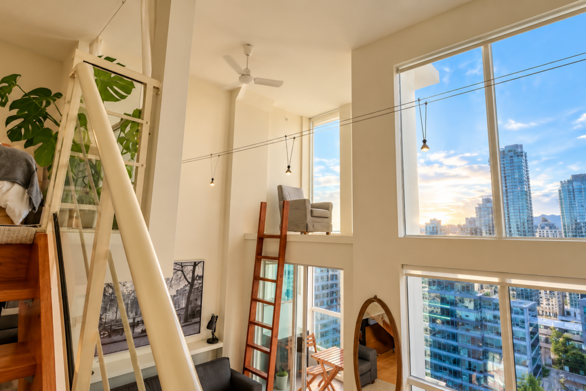 Bright modern loft with large windows and city views, featuring a wooden ladder, plant, and bed nook on the left side.