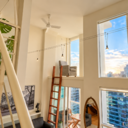 Bright modern loft with large windows and city views, featuring a wooden ladder, plant, and bed nook on the left side.