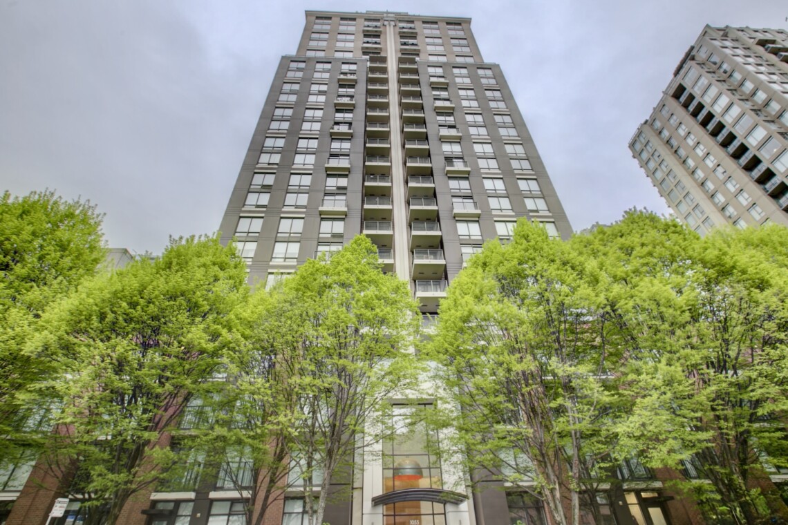 Tall modern residential high-rise viewed from street level, with green trees at the bottom and a blue sky above.