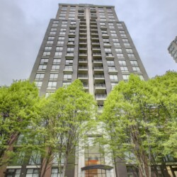 Tall modern residential high-rise viewed from street level, with green trees at the bottom and a blue sky above.