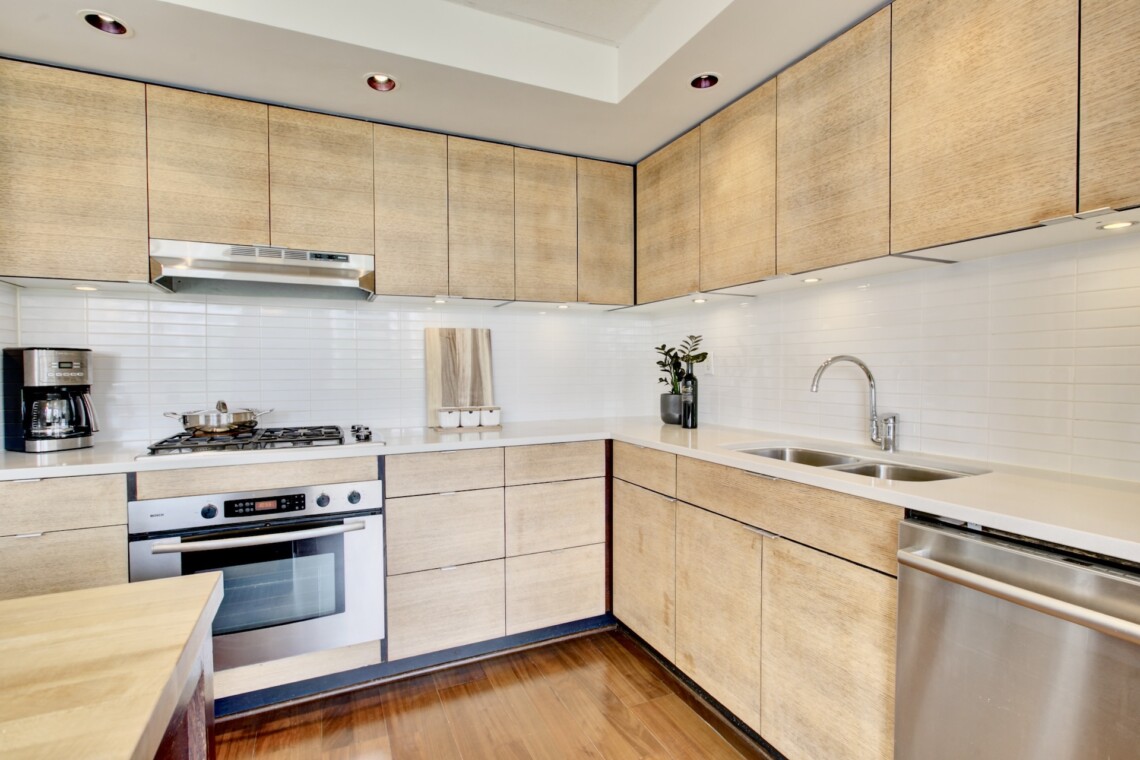 Modern L-shaped kitchen with light wood cabinets, white tile backsplash, and stainless steel appliances including oven, stove, and dishwasher