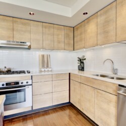 Modern L-shaped kitchen with light wood cabinets, white tile backsplash, and stainless steel appliances including oven, stove, and dishwasher