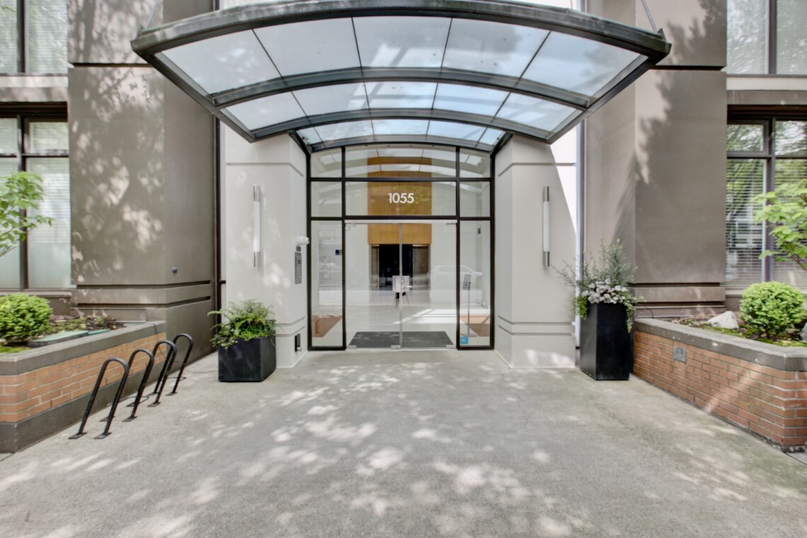 Front entrance of a modern building with a curved glass canopy, double doors, and the number 1055 above the doorway.