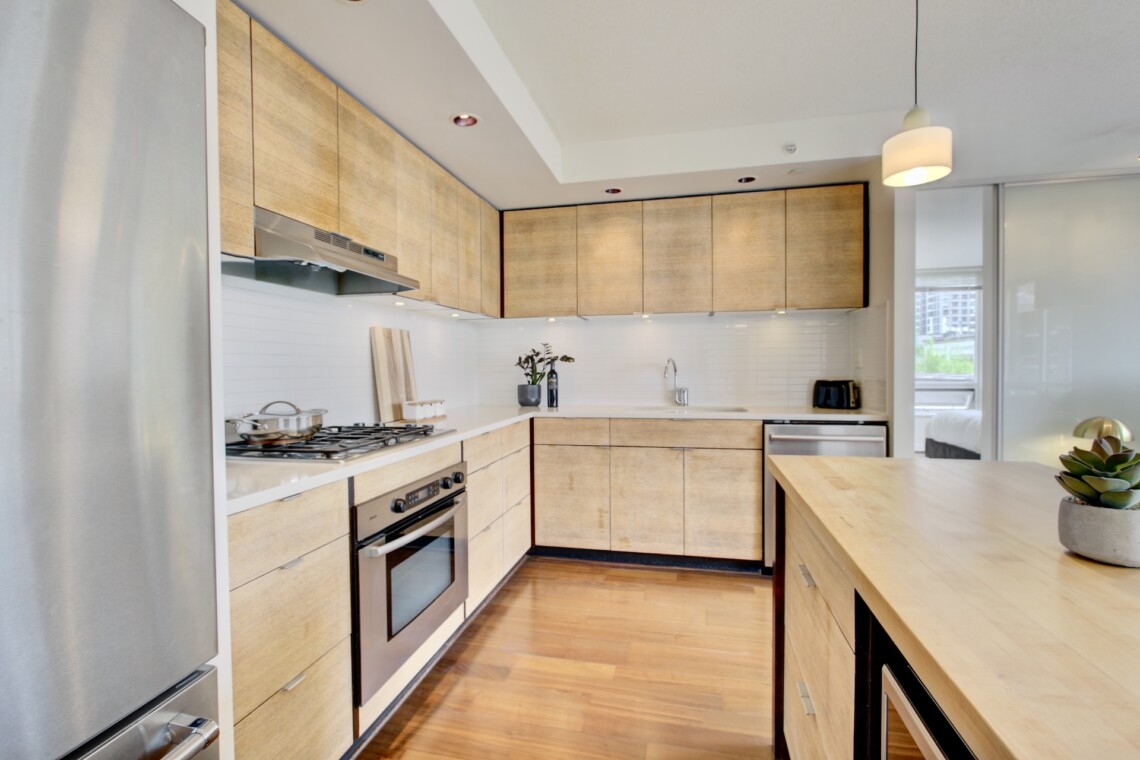 Modern U-shaped kitchen with light wood cabinets, stainless steel appliances, and white backsplash; island in foreground and pendant light nearby.