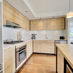 Modern U-shaped kitchen with light wood cabinets, stainless steel appliances, and white backsplash; island in foreground and pendant light nearby.