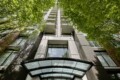 Low-angle view of a tall modern apartment building framed by green trees, with a glass entrance canopy at the bottom.