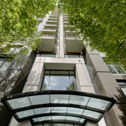 Low-angle view of a tall modern apartment building framed by green trees, with a glass entrance canopy at the bottom.