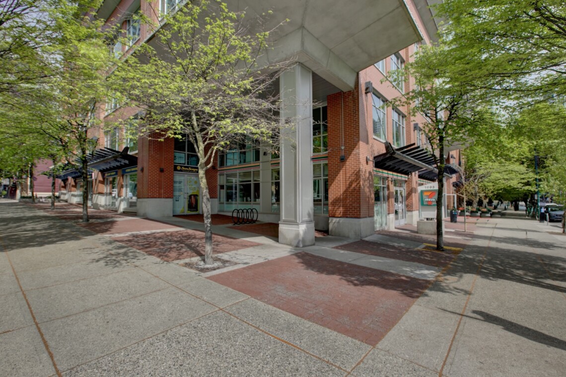 Brick building with large windows along a tree-lined sidewalk; a 7-Eleven sign visible on the corner of the street.