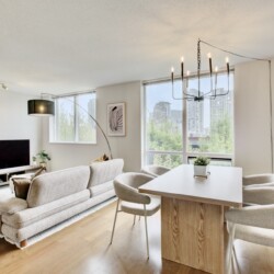 Bright, modern living room with a beige sofa, TV console, and large windows showing a city view.