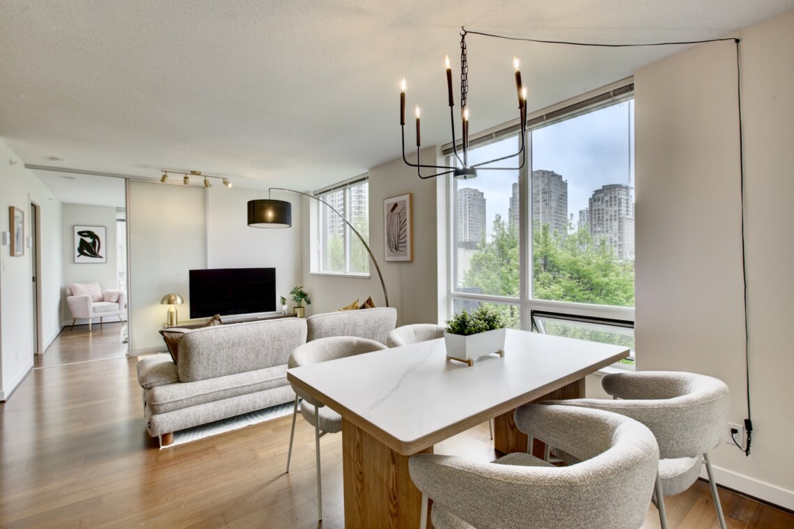 Bright open-concept living room with a white dining table and beige chairs, overlooking a cityscape through large windows.