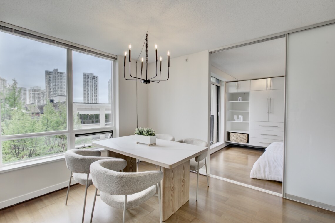 Modern dining area with a light wood table, soft gray chairs, and a black multi-arm chandelier, near large windows overlooking the city.