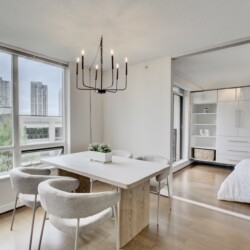 Modern dining area with a light wood table, soft gray chairs, and a black multi-arm chandelier, near large windows overlooking the city.