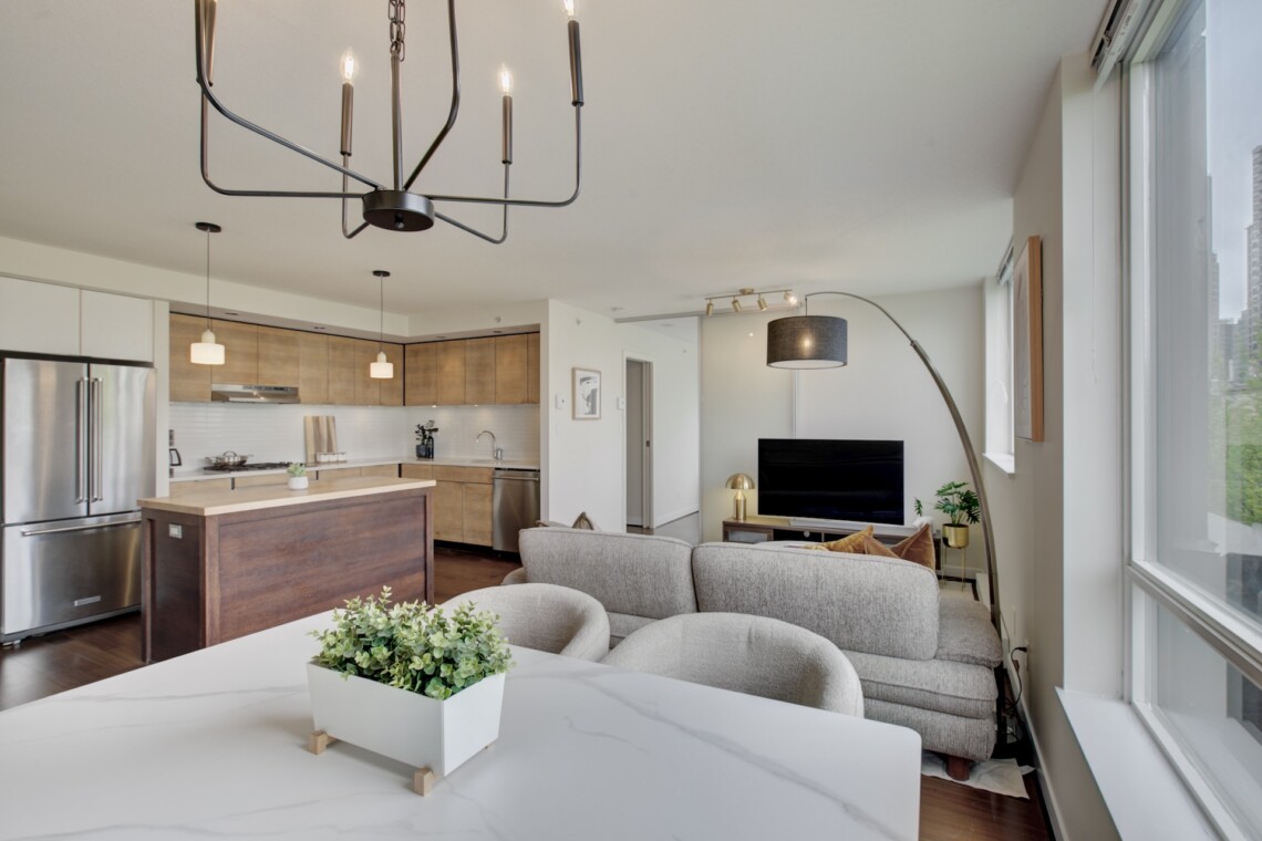Bright open-plan living area with a kitchen on the left, a large black metal chandelier, gray sofa, and big windows in the background.