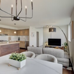 Bright open-plan living area with a kitchen on the left, a large black metal chandelier, gray sofa, and big windows in the background.