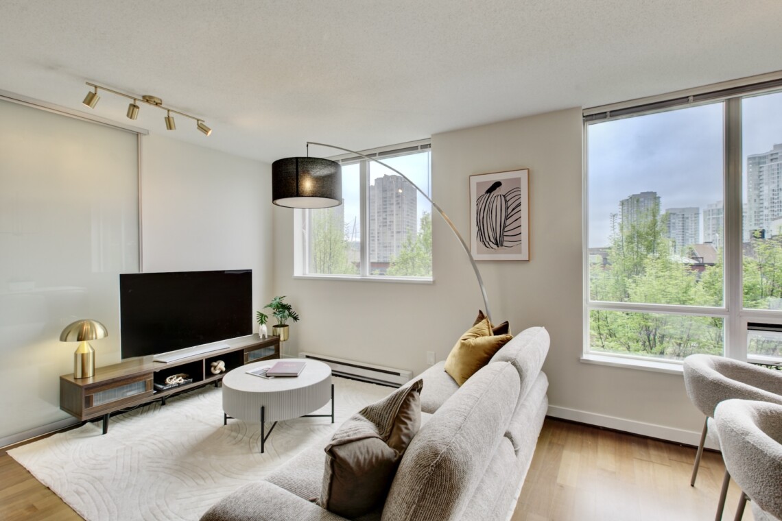 Bright living room with large windows overlooking the city, a gray sofa with mustard cushions, a round coffee table, and a TV on a wooden console.