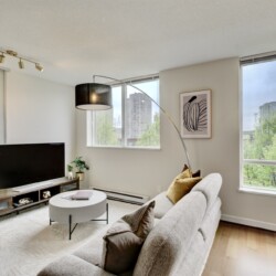 Bright living room with large windows overlooking the city, a gray sofa with mustard cushions, a round coffee table, and a TV on a wooden console.