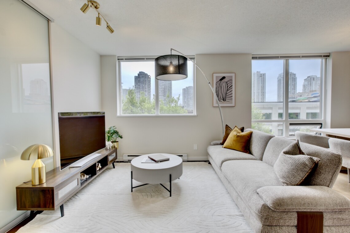 Bright living room with a beige sectional, round coffee table, and a wall of windows overlooking a city skyline.