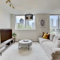 Bright living room with a beige sectional, round coffee table, and a wall of windows overlooking a city skyline.
