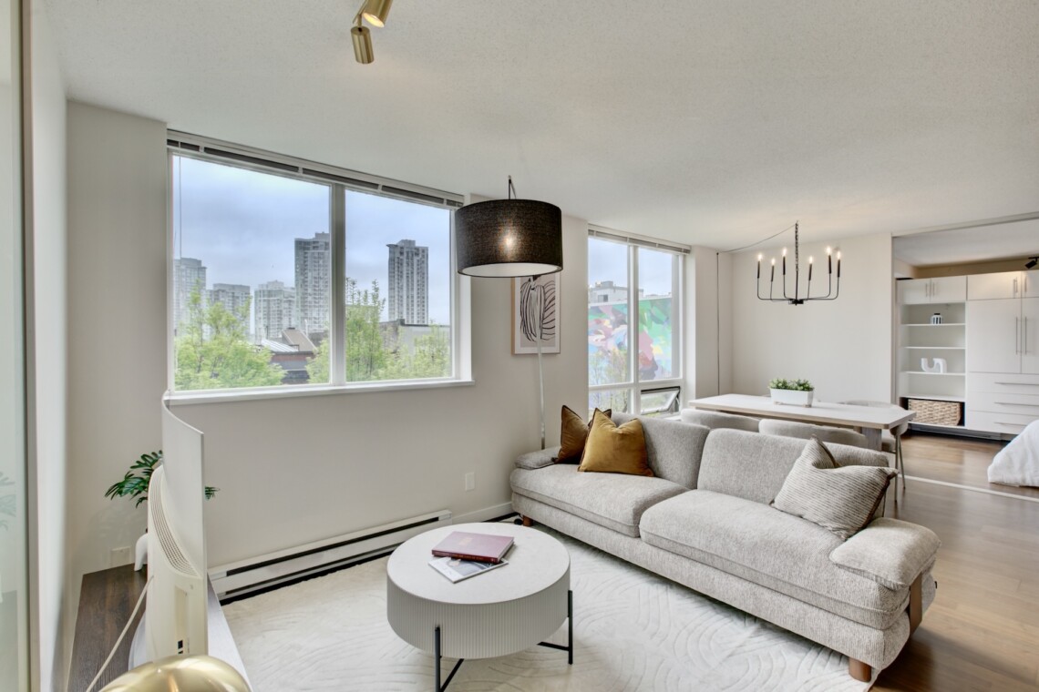 Bright open-plan living area with a gray sectional sofa, round white coffee table, and large windows showing a city skyline; dining area and white cabinetry visible in the background.