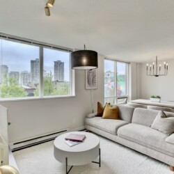 Bright open-plan living area with a gray sectional sofa, round white coffee table, and large windows showing a city skyline; dining area and white cabinetry visible in the background.