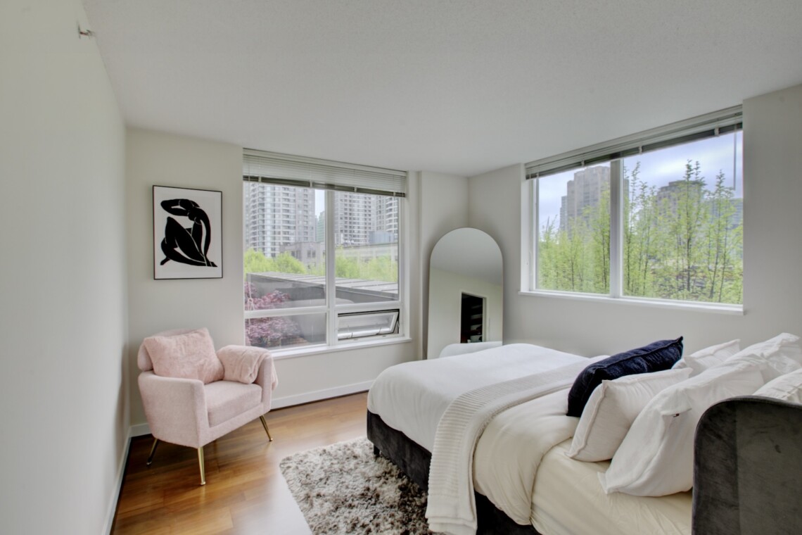 Bright bedroom with white bedding, a pink chair, and large windows showing city buildings outside.