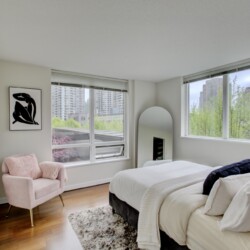 Bright bedroom with white bedding, a pink chair, and large windows showing city buildings outside.