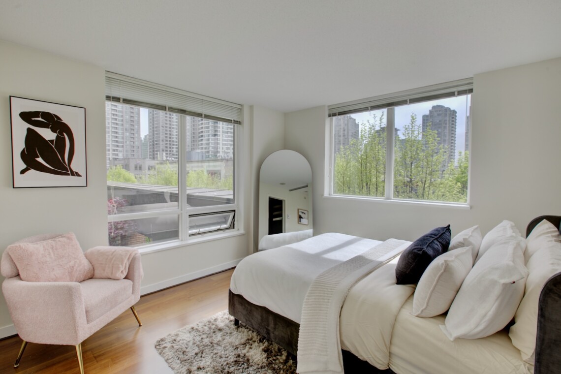 Bright modern bedroom with a white bed, pink accent chair, and large windows showing a city skyline and greenery outside.