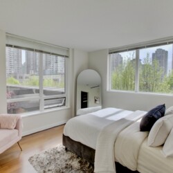 Bright modern bedroom with a white bed, pink accent chair, and large windows showing a city skyline and greenery outside.
