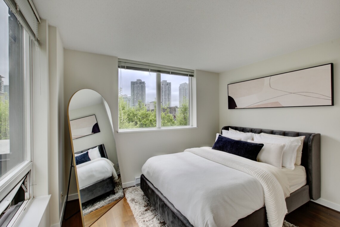 Bright modern bedroom with a gray upholstered bed, white bedding, and a large window showing a city skyline.