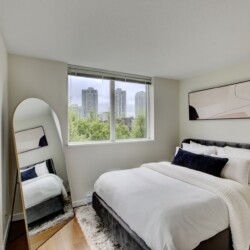 Bright modern bedroom with a gray upholstered bed, white bedding, and a large window showing a city skyline.